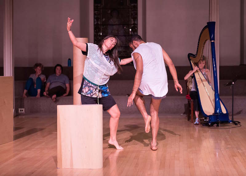 Wooden triangles and tubes are scatted. Two dancers stand around them. One is balancing on one leg